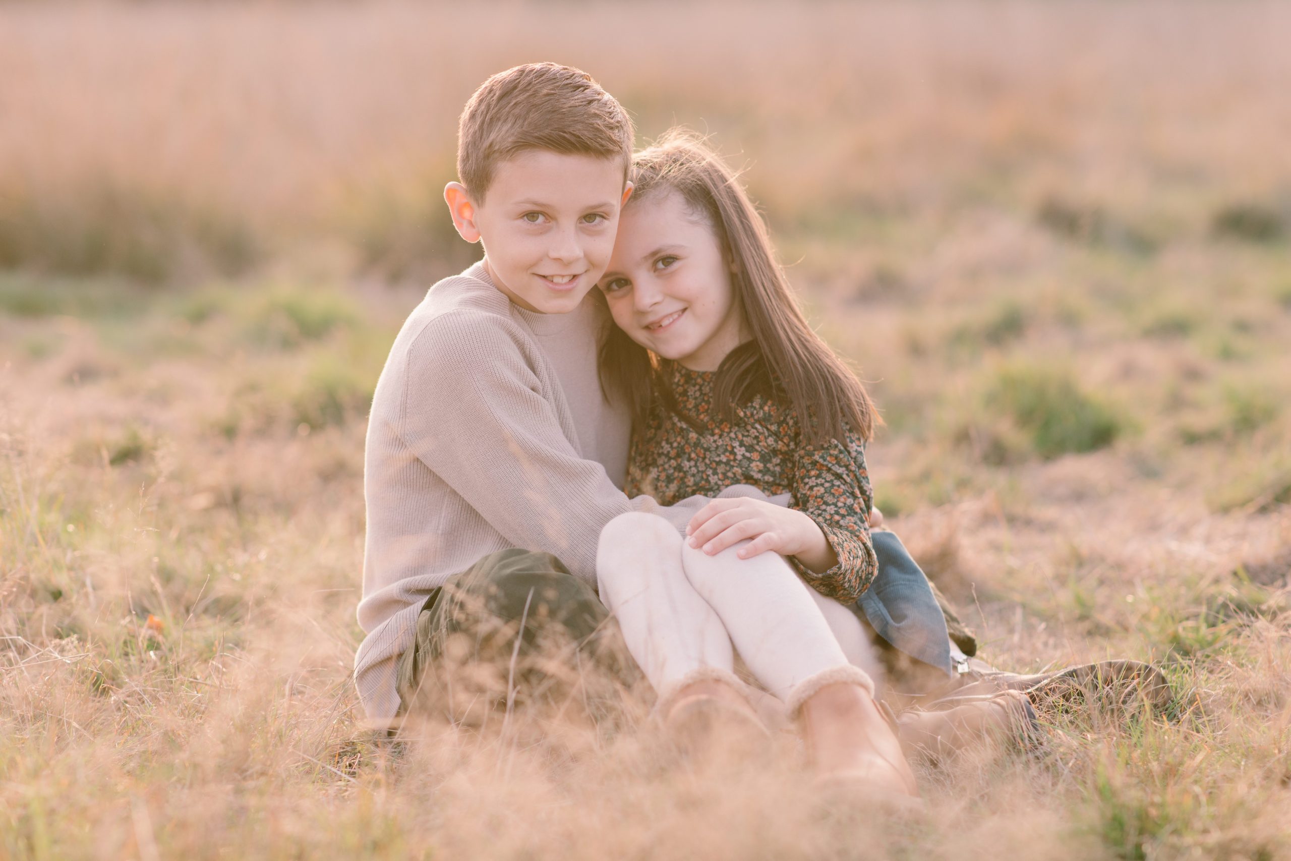 Young brother and sister sitting down in the grass looking at the camera and cuddling - Hampshire Family Photographer 